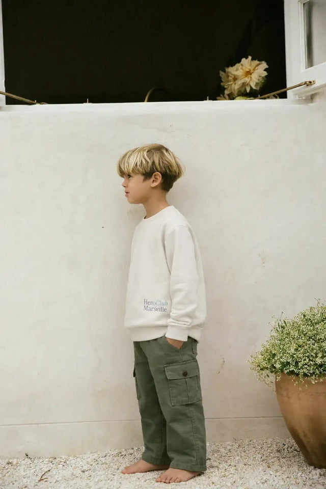Un enfant portant un sweat-shirt blanc doux à col rond avec ’heroclub marseille’ imprimé en texte gris subtil sur la poitrine gauche.