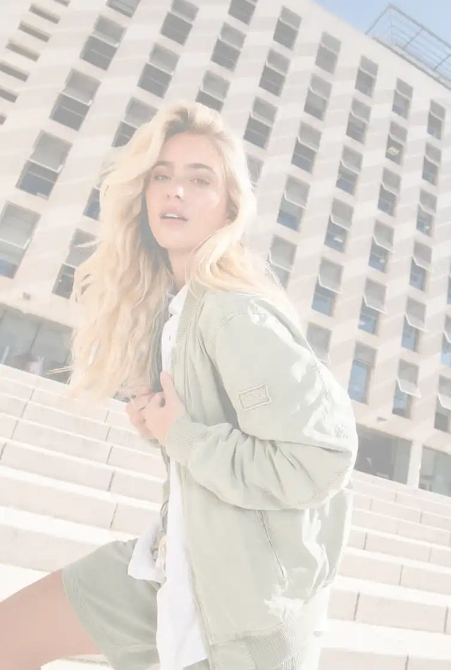 Une femme aux cheveux longs et ondulés blonds portant une veste bomber vert clair et un short assorti sur un t-shirt blanc.